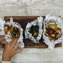Dziecięce rączki sięgają po pieczone warzywa w folii. Idealne co na ognisko do jedzenia: ziemniaki, marchewka, cebula i czosnek.
