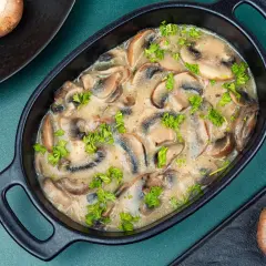 Wykwintny sos pieczarkowy w czarnej miseczce, udekorowany natką pietruszki. Obok leżą pieczarki.
