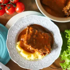 Pyszny sos z żeberek podany z purée ziemniaczanym na ozdobnym talerzu. Obok świeża sałata i pomidory.