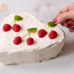 Jak udekorować tort owocami? Ręka z malinowymi paznokciami dodaje listek mięty do sercowatego tortu z malinami.