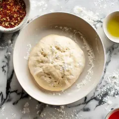 Ciasto na pinsę w misce, obok miseczka z płatkami chili, oliwą i sosami. Przepis na ciasto czeka!
