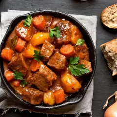 Gulasz wołowy z marchewką i ziemniakami w żeliwnej patelni. Ile kostek rosołowych na litr wody? Obok cebula, marchewka i chleb.