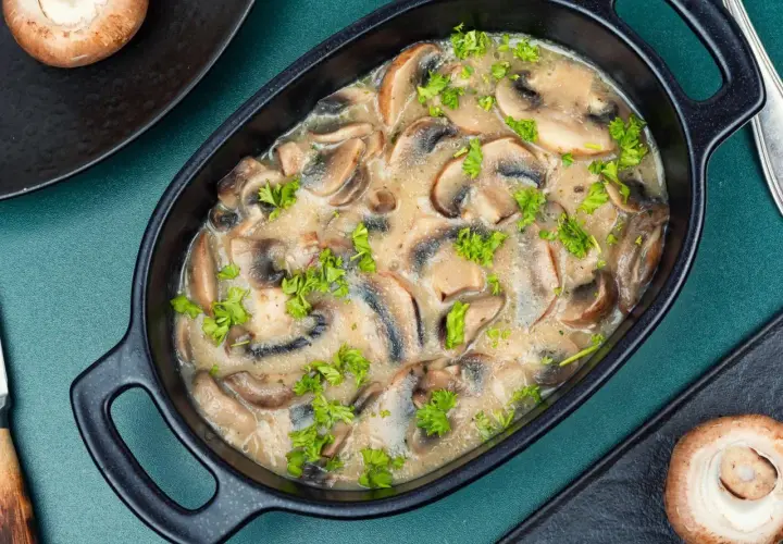 Wykwintny sos pieczarkowy w czarnej miseczce, udekorowany natką pietruszki. Obok leżą pieczarki.