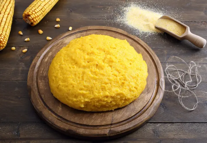 Puszysta polenta z mąki kukurydzianej na drewnianej desce, obok kolby kukurydzy i garści mąki.