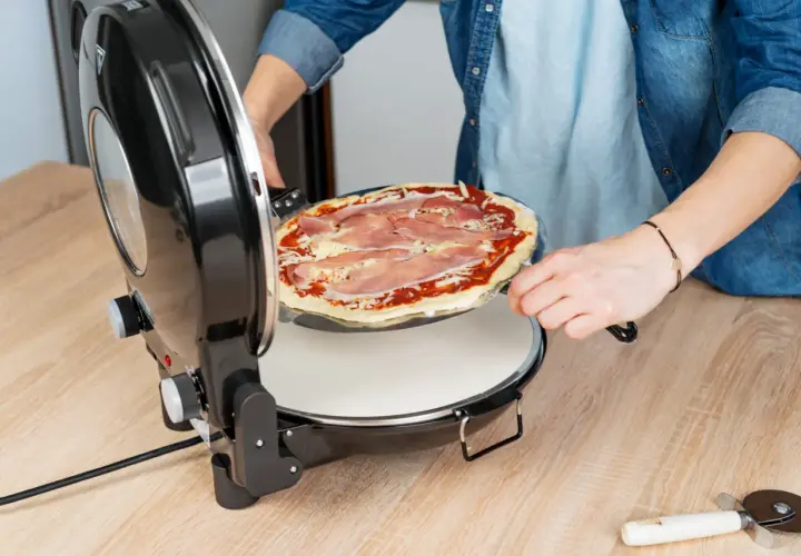 Osoba wkłada do pieca pizzę z szynką i serem. Już niedługo będzie można cieszyć się smakiem domowej pizzy.