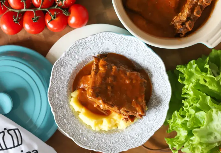 Pyszny sos z żeberek podany z purée ziemniaczanym na ozdobnym talerzu. Obok świeża sałata i pomidory.