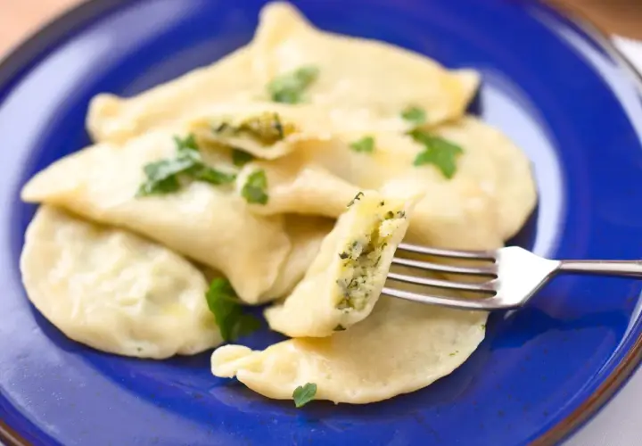 Pyszne pierogi z farszem ze szpinaku, udekorowane natką pietruszki, prezentowane na niebieskim talerzu. Widoczny widelec pokazuje bogate wnętrze.