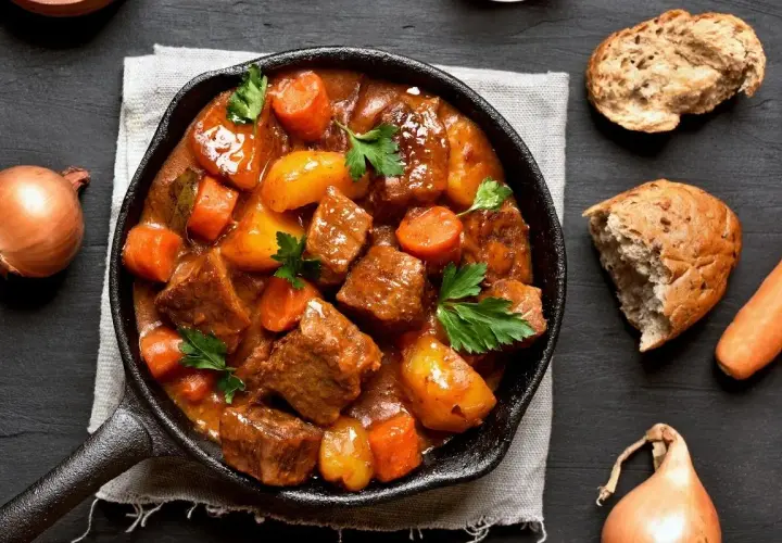 Gulasz wołowy z marchewką i ziemniakami w żeliwnej patelni. Ile kostek rosołowych na litr wody? Obok cebula, marchewka i chleb.