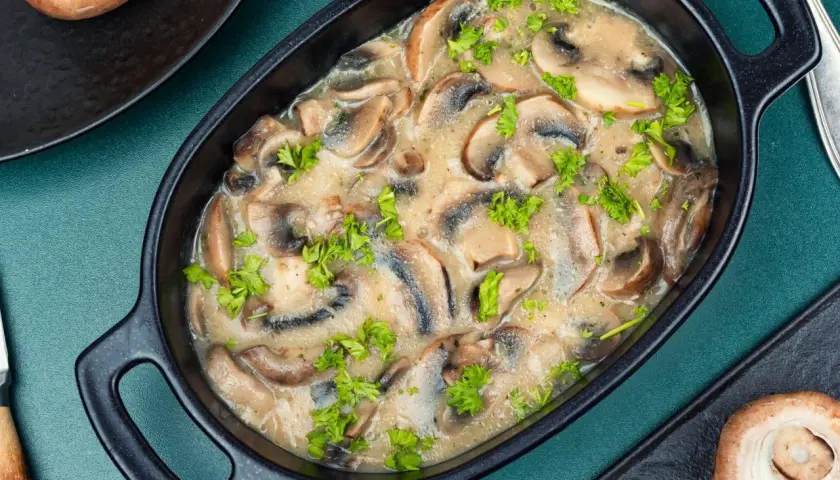 Wykwintny sos pieczarkowy w czarnej miseczce, udekorowany natką pietruszki. Obok leżą pieczarki.
