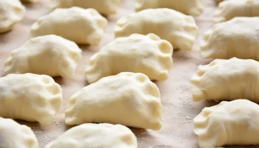 Surowe ciasto na pierogi z olejem, uformowane w kształt półksiężyców, leży na blacie posypanym mąką.