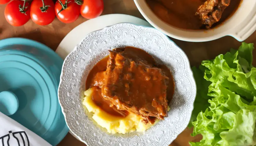 Pyszny sos z żeberek podany z purée ziemniaczanym na ozdobnym talerzu. Obok świeża sałata i pomidory.