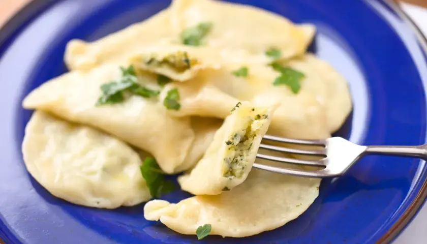 Pyszne pierogi z farszem ze szpinaku, udekorowane natką pietruszki, prezentowane na niebieskim talerzu. Widoczny widelec pokazuje bogate wnętrze.