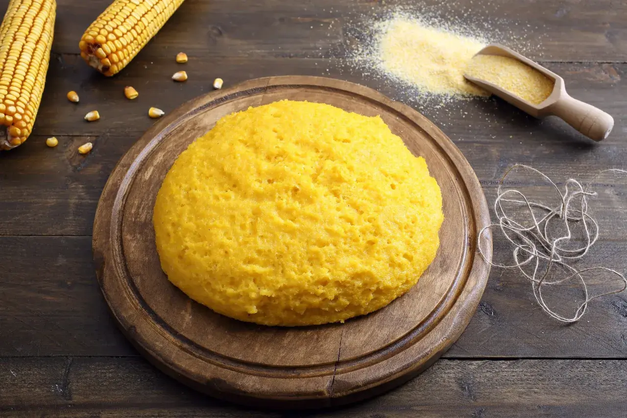 Puszysta polenta z mąki kukurydzianej na drewnianej desce, obok kolby kukurydzy i garści mąki.