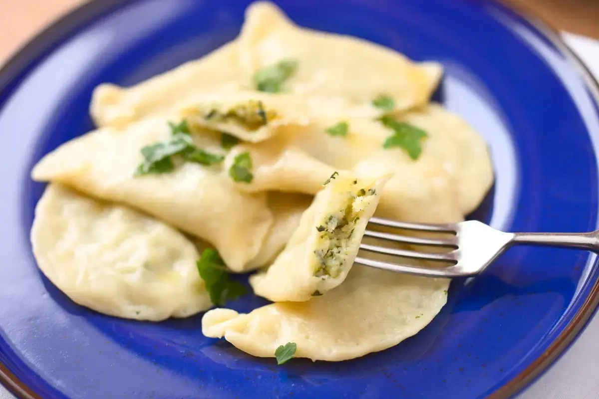 Pyszne pierogi z farszem ze szpinaku, udekorowane natką pietruszki, prezentowane na niebieskim talerzu. Widoczny widelec pokazuje bogate wnętrze.