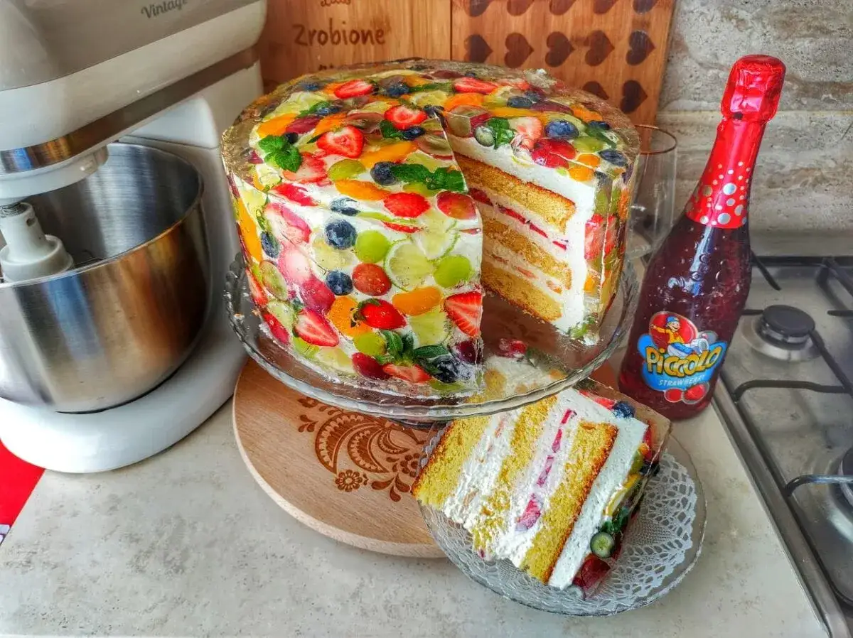 Jak udekorować tort owocami? Kolorowy tort z galaretką i owocami, obok stoi butelka Piccolo.