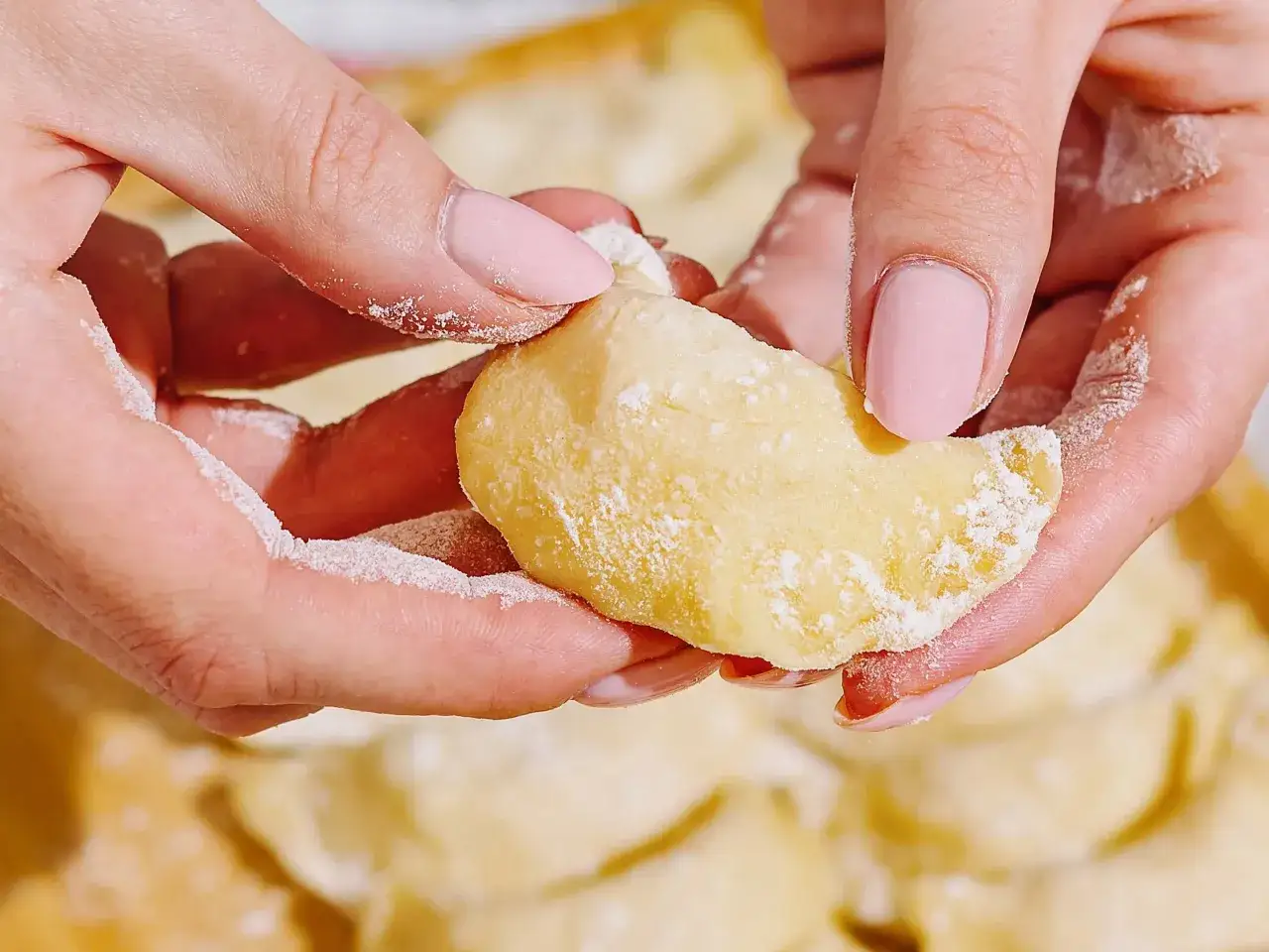 Dłonie formują ciasto na pierogi z olejem, posypane mąką. W tle widać gotowe pierogi.
