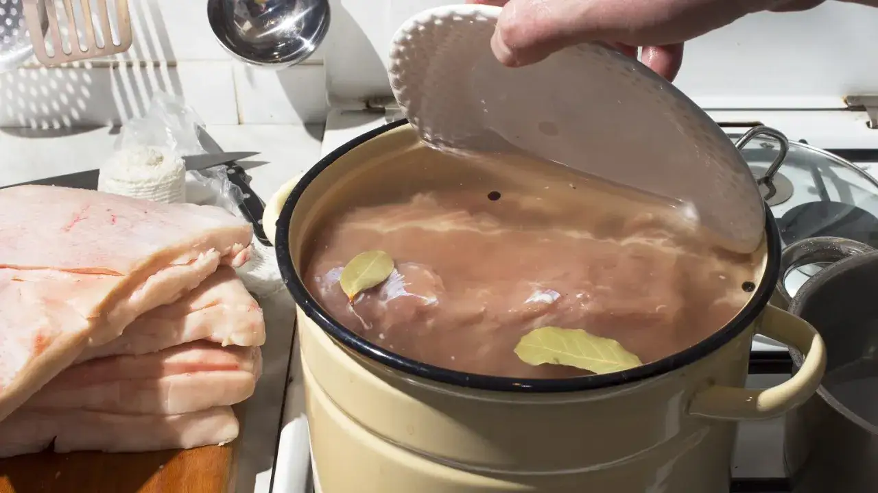 Mięso w garnku z zalewą, przygotowywane do peklowania. Obok stos surowego boczku.