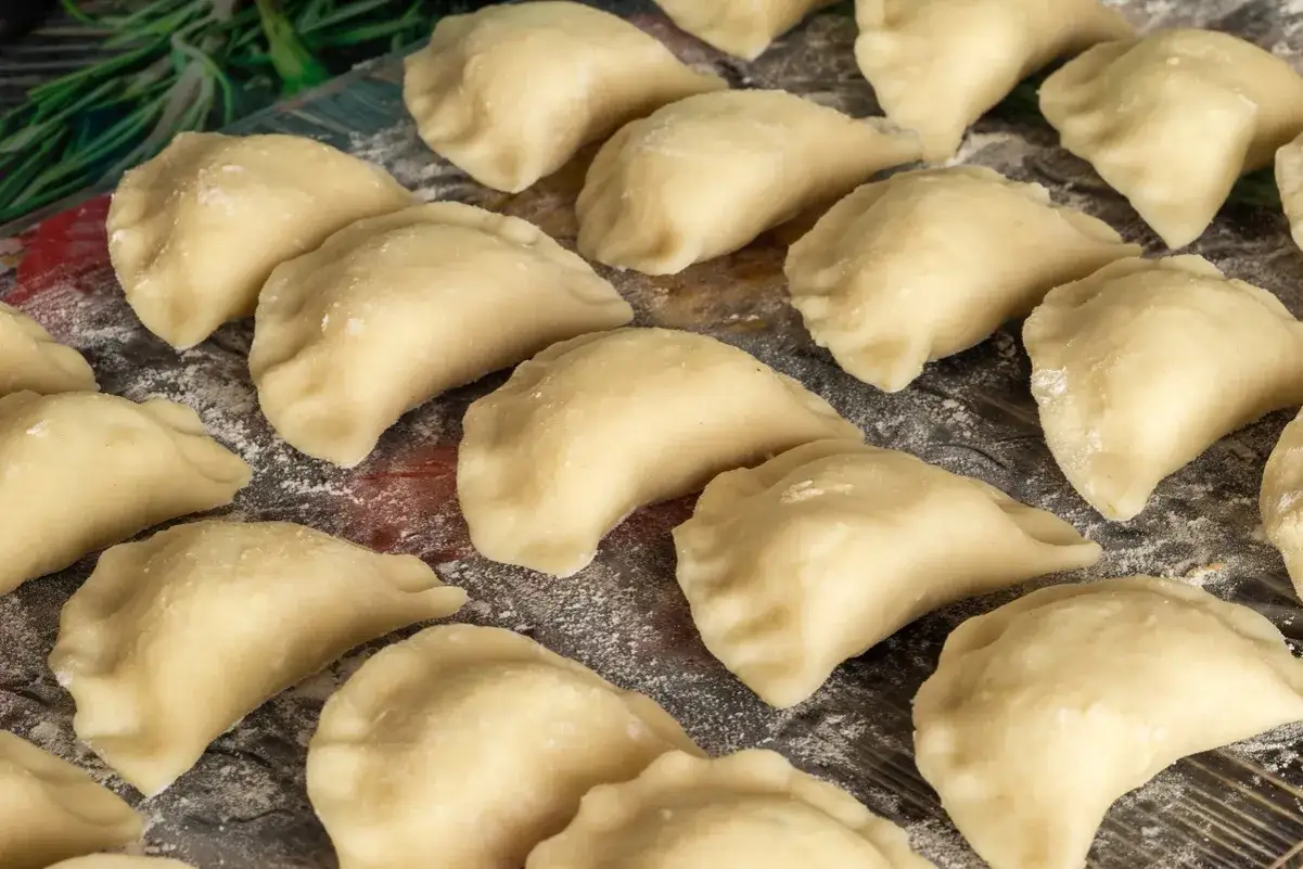 Surowe ciasto na pierogi, zrobione z dodatkiem oleju, czeka na nadzienie. Widać je na drewnianym blacie, posypanym mąką.