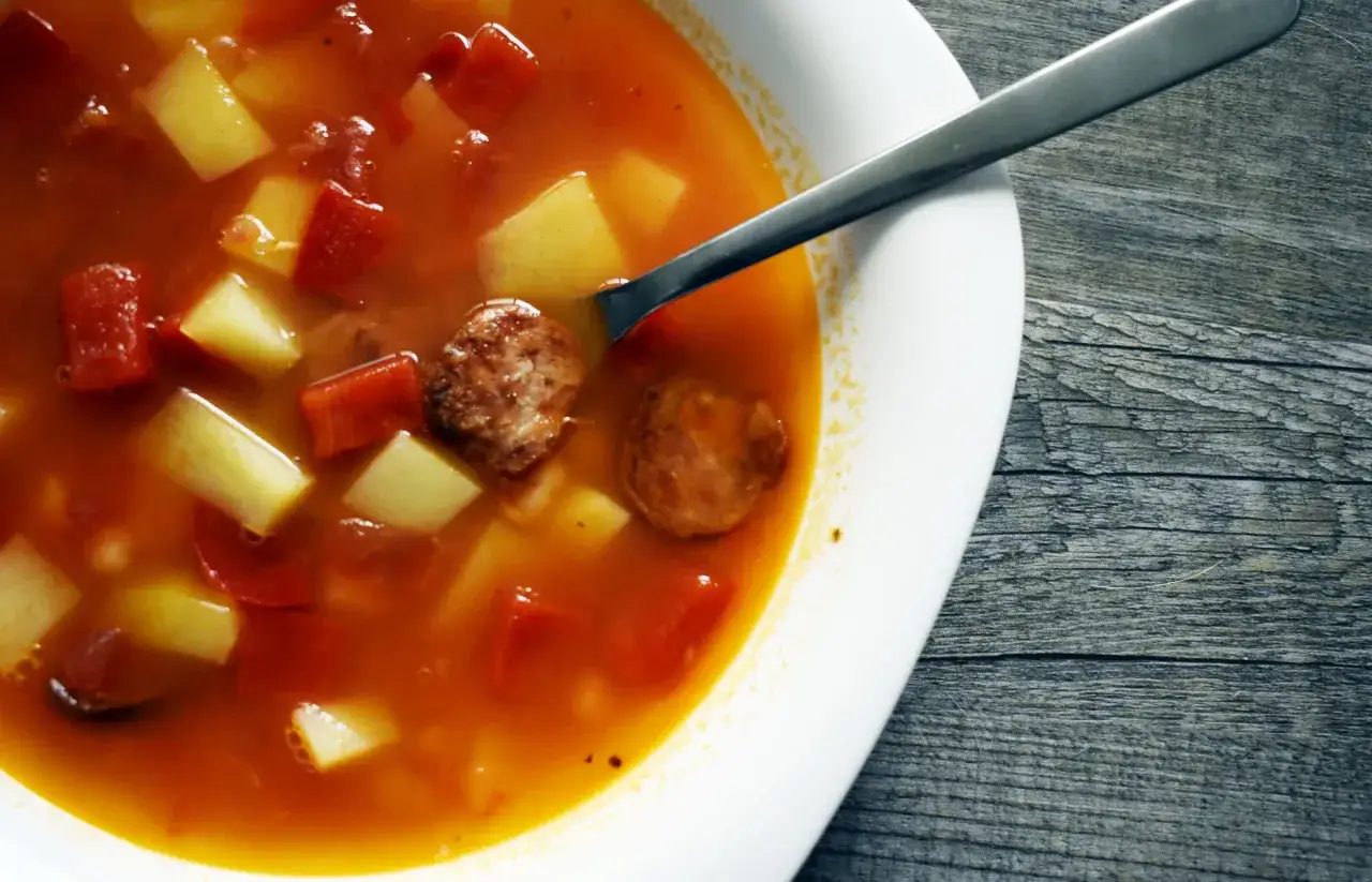 Prosta zupa paprykowa z kawałkami ziemniaków i mięsa, podana w białej misce z metalową łyżką na drewnianym tle.
