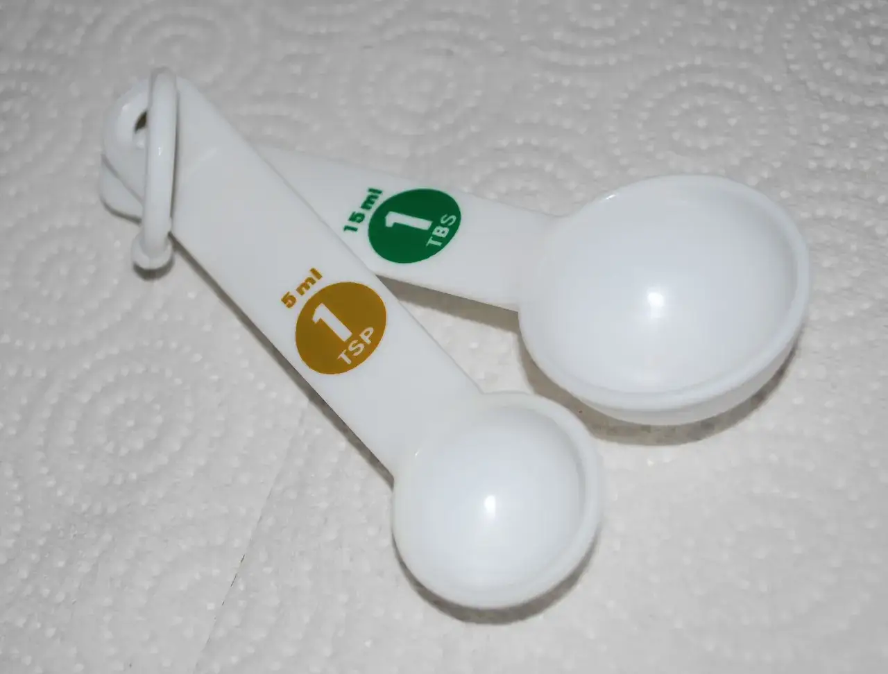 Dwie białe łyżki miarowe: jedna 5 ml (1 łyżeczka), druga 15 ml (1 łyżka stołowa).