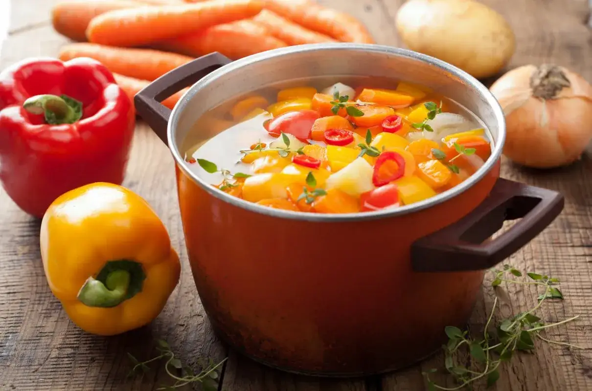 Gotuje się prosta zupa paprykowa. W garnku bulgoczą warzywa: papryka, marchewka, ziemniaki. Obok leżą świeże papryki, marchewki i cebula.