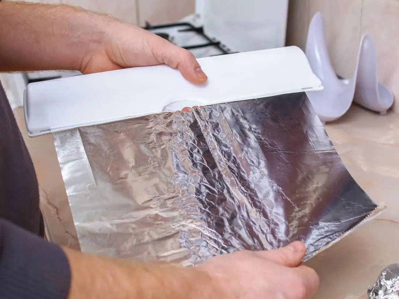Ręce trzymają folię aluminiową z dozownika. Czy można użyć folii aluminiowej zamiast papieru do pieczenia? Tak, to świetna alternatywa.